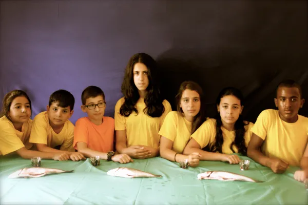 Photo of seven children sitting behind a table with turquoise tablecloths, where there are three fishes and four glasses. All children wear yellow t-shirts and their right arm is resting on the table, except the third from the left, which T-Shirt is orange and the fourth person, in the middle, which is also the tallest, has long black hair and has both hands resting on the table.